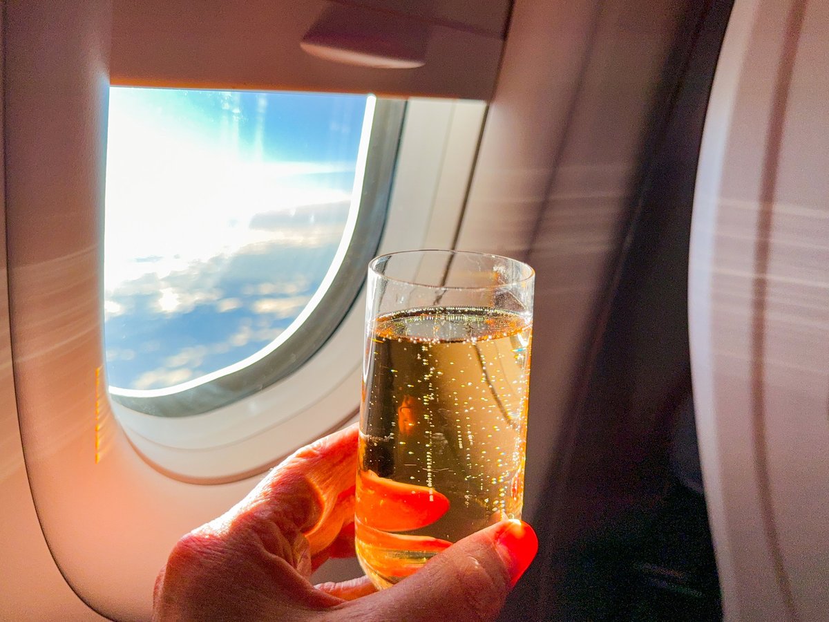Person holding a glass of champagne in front of a window in the business class cabin of a passenger jet